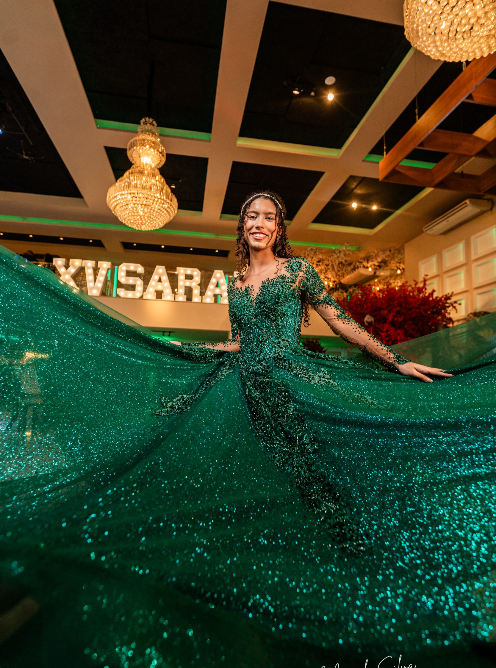 Debutante em vestido verde bordado abrindo a saia sob letreiro luminoso XV Sara e lustres de cristal