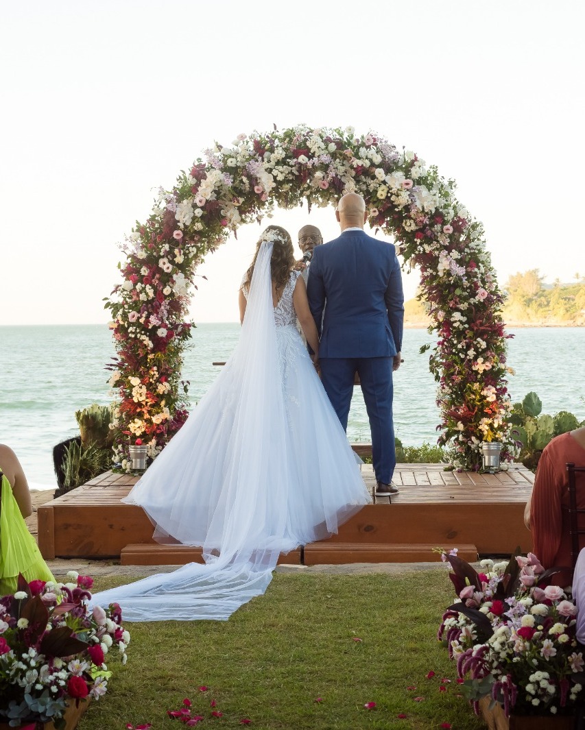 Noivos de costas sob arco de flores durante cerimônia de casamento à beira-mar